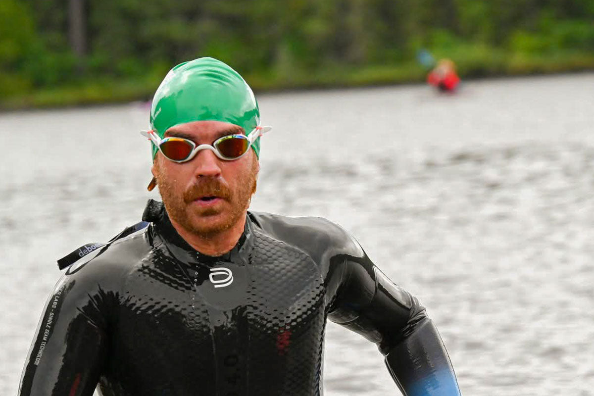 Man in a wetsuit and green swim cap runs out of the water toward transition.