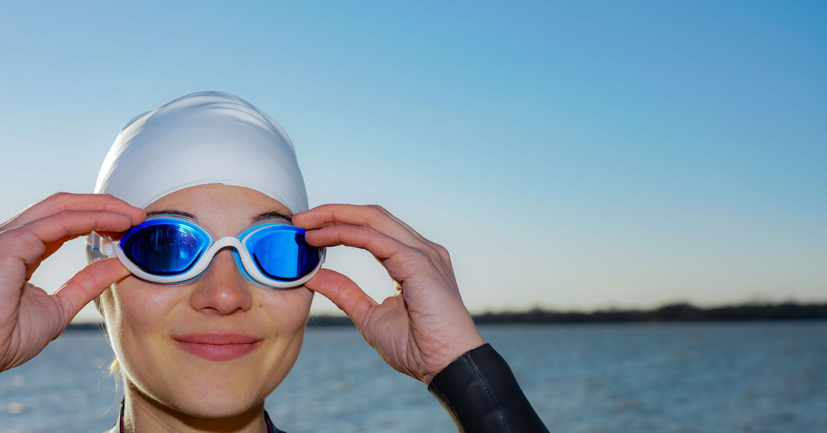 A triathlete in a wetsuit near a lake wears blue tinted Snake & Pig's comfortable swim goggles and a white swim cap