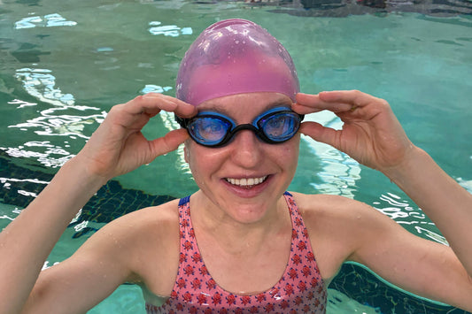 Woman standing in a swimming pool wearing black clear Basilisk goggles and a pink cap. She is touching the sides of the goggles with her hands.