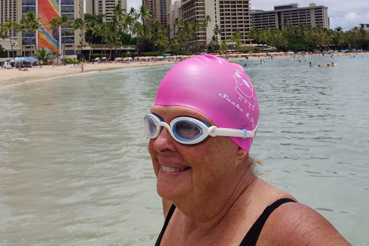 A woman in clear Snake & Pig goggles and a pink swim cap stand off the shore in Waikiki