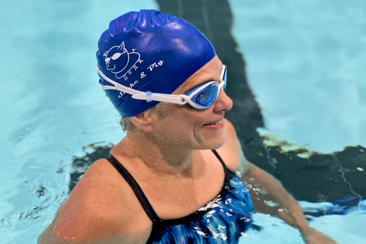 Woman standing in a pool facing right to show the side of the light blue lens goggles. She is wearing a blue Snake & Pig swim cap and a blue and black striped swimsuit.