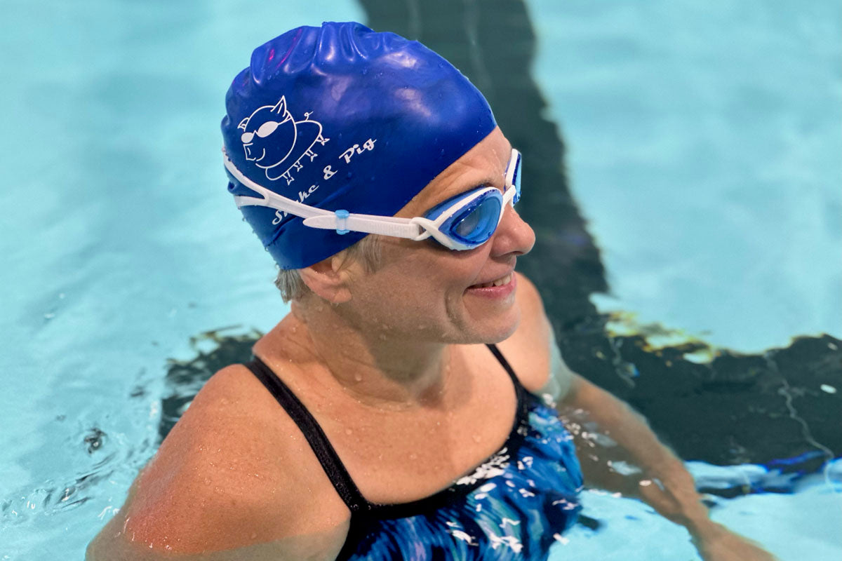 Woman standing in a pool facing right to show the side of the light blue lens goggles. She is wearing a blue Snake & Pig swim cap and a blue and black striped swimsuit.