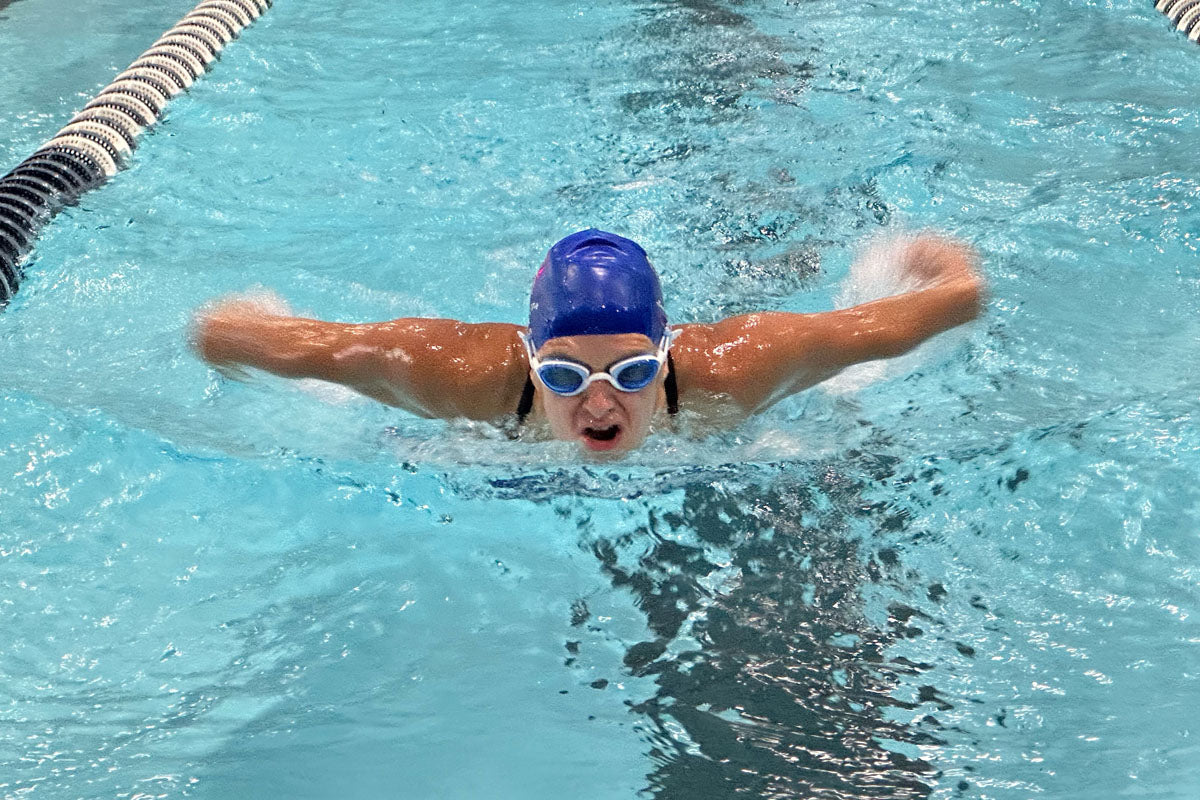 A woman wearing light blue lens goggles swims butterfly in a swimming pool.