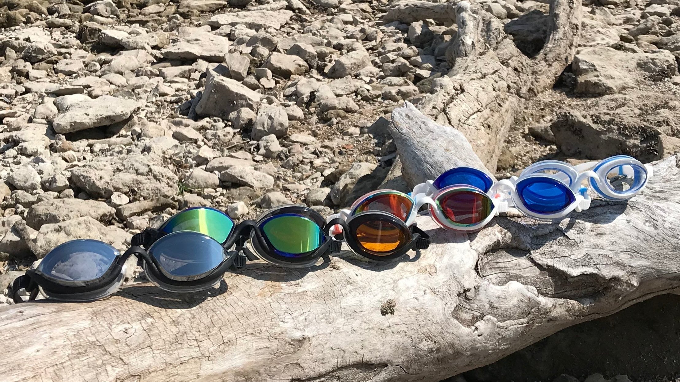 Six of the seven tints of comfortable swim goggles offered, sitting on a piece of driftwood on a rocky shoreline