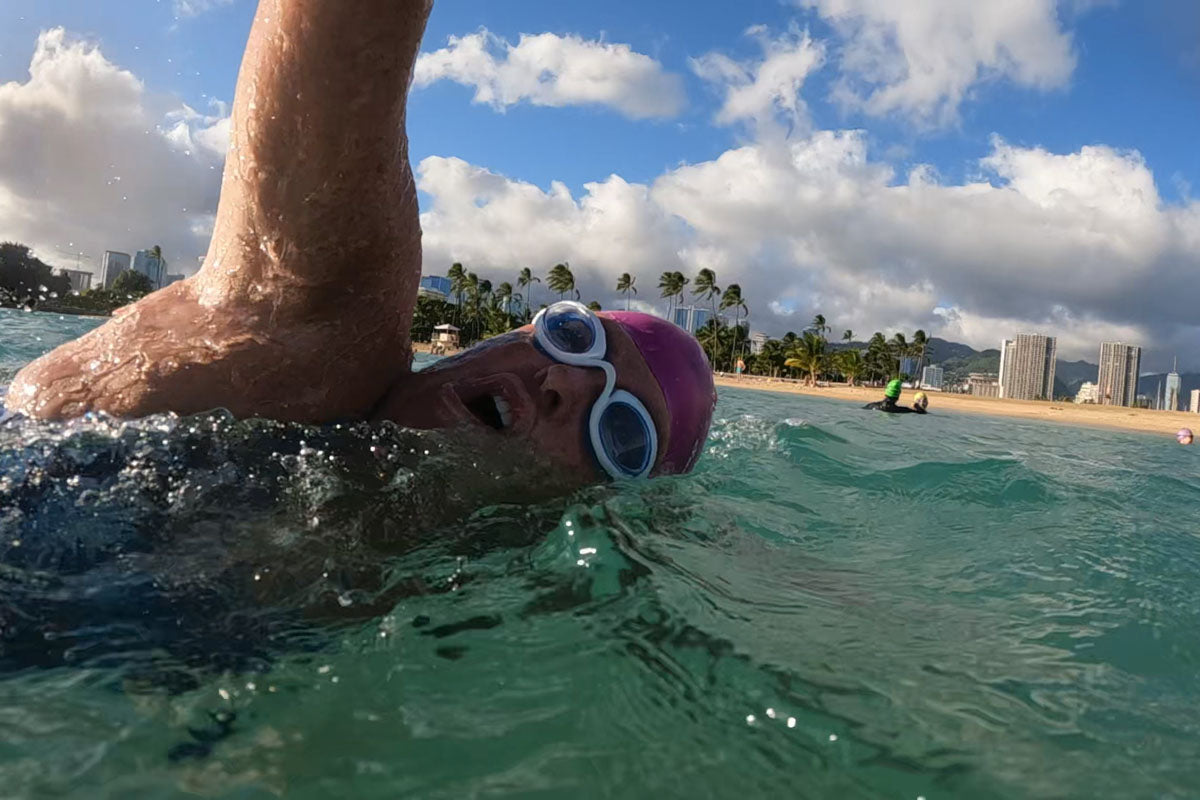 A woman takes a breath to the side while swimming freestyle in the ocean. She is wearing blue clear Snake & Pig goggles and a pink swim cap.