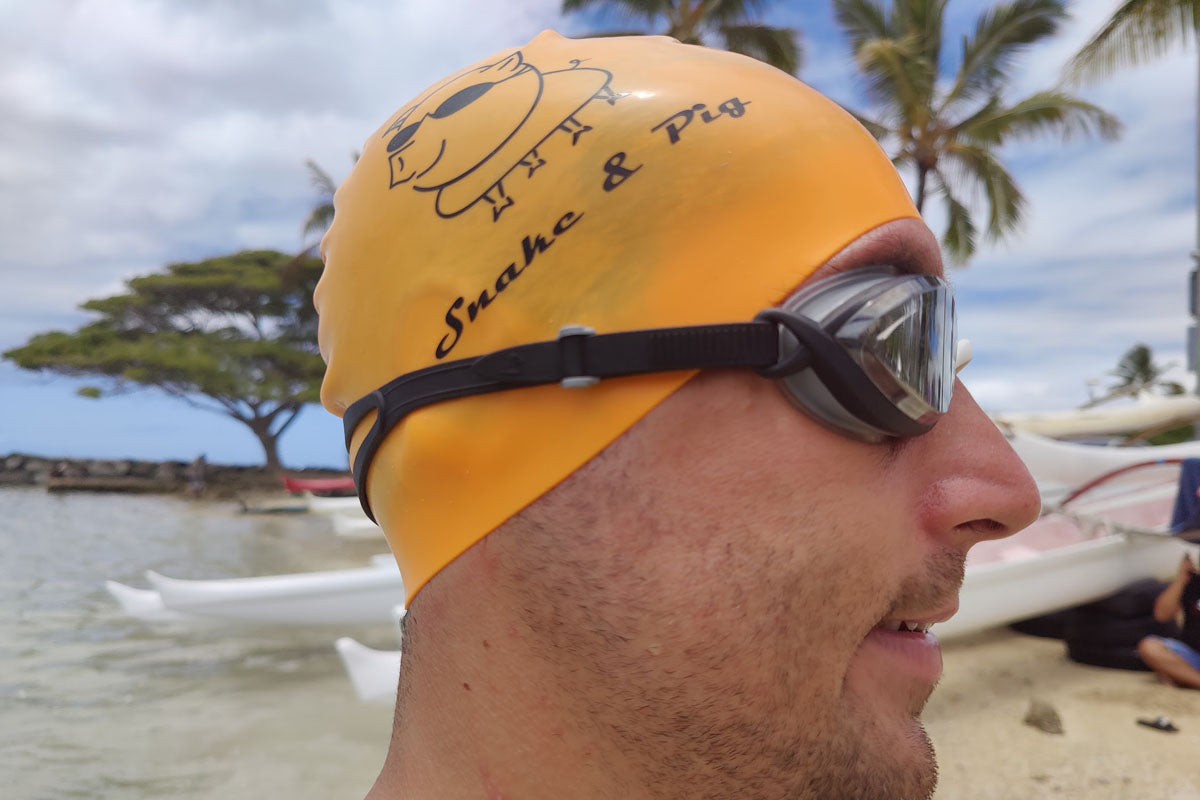 Man on the beach wearing an orange Snake & Pig swim cap and black clear, metallized goggles