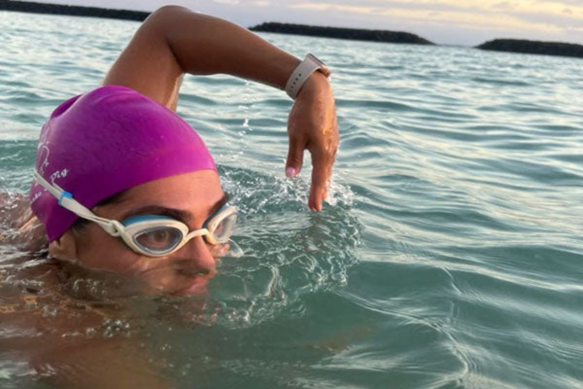 A woman in the ocean sights above the water while wearing a purple swim cap and clear Snake & Pig goggles