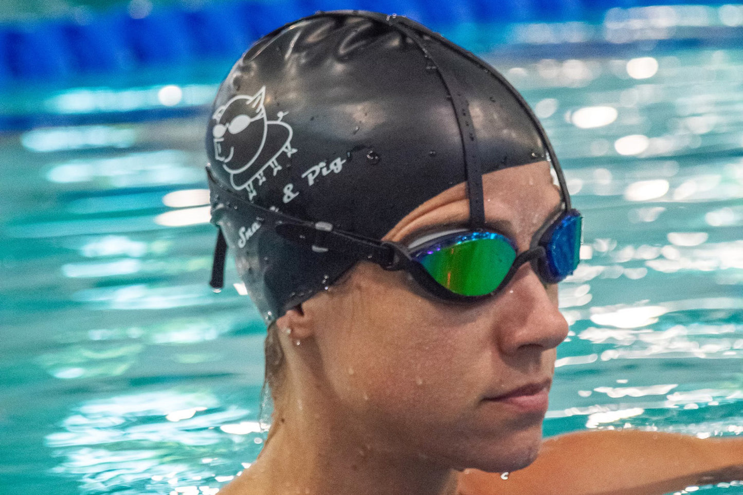 A swimmer in a pool wears a calm expression, a black swim cap with a Snake & Pig logo, and a pair of top anchor goggles with a dark tint