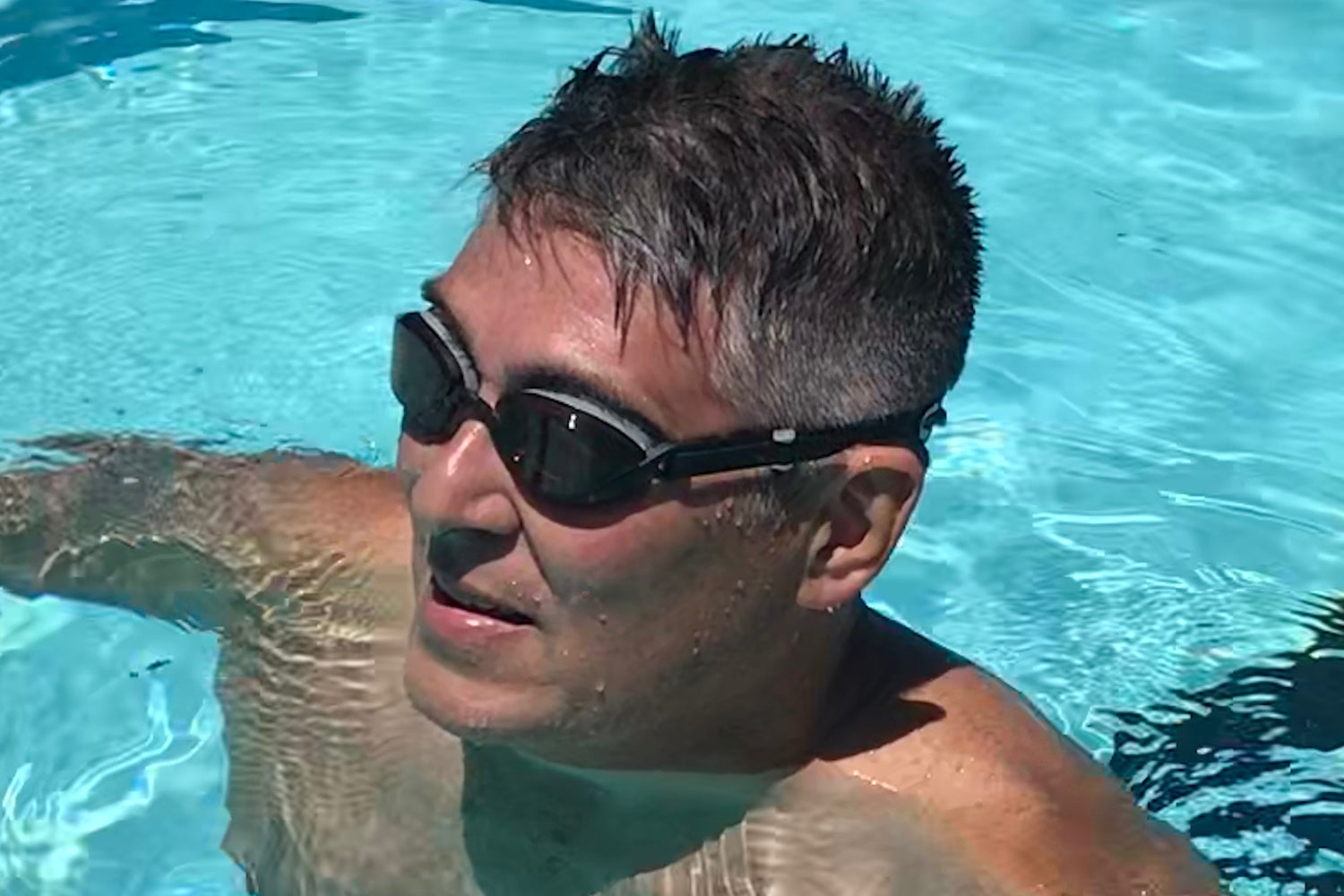 A man in a swimming pool wears amber goggles in bright sunlight