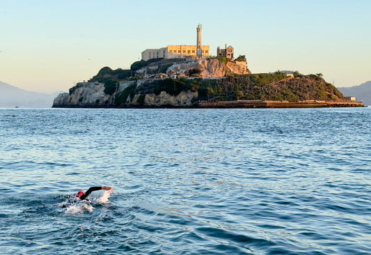 A swimmer in a wetsuit and pink swim cap swims toward Alcatraz Island on a clear, sunny day.