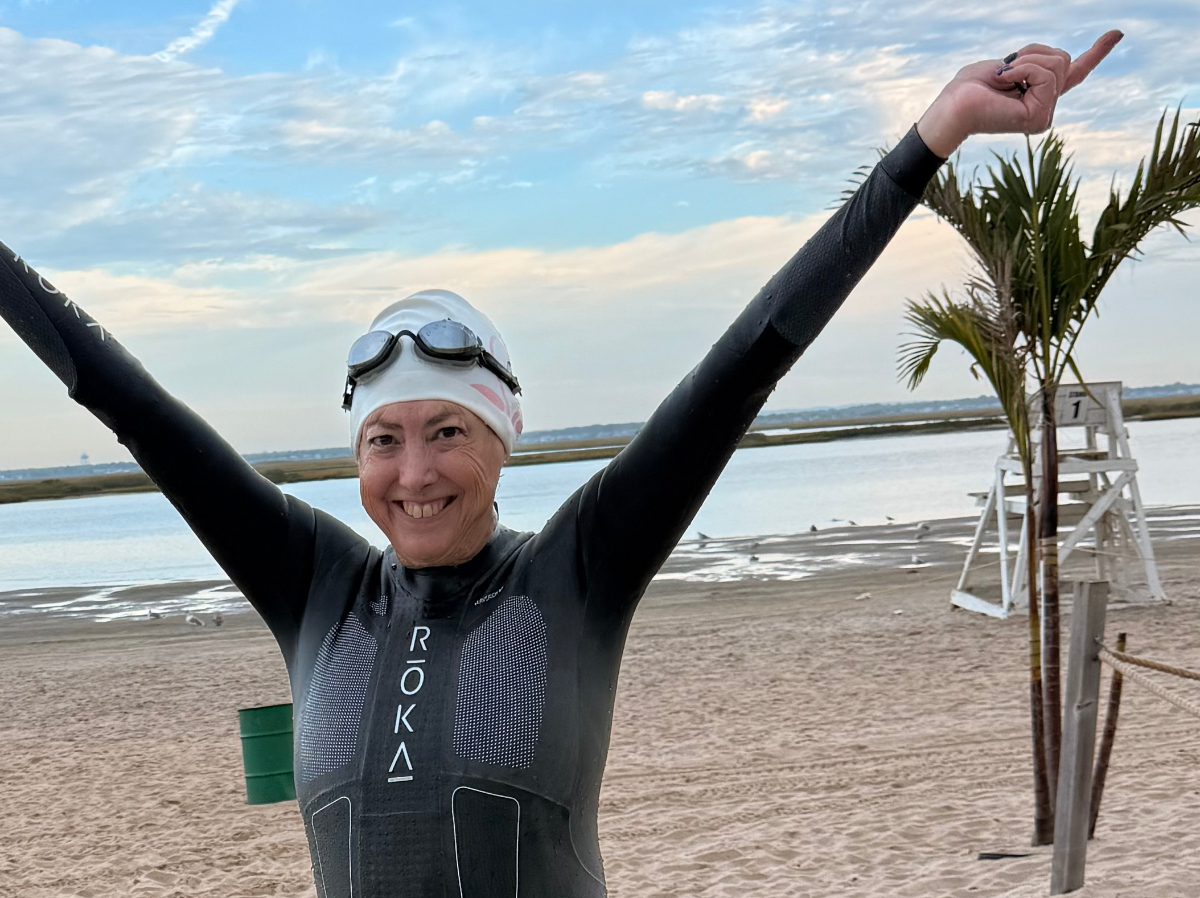 Hilary Topper wearing black clear metallized goggles, white cap and wetsuit, arms outstretched, on a beach