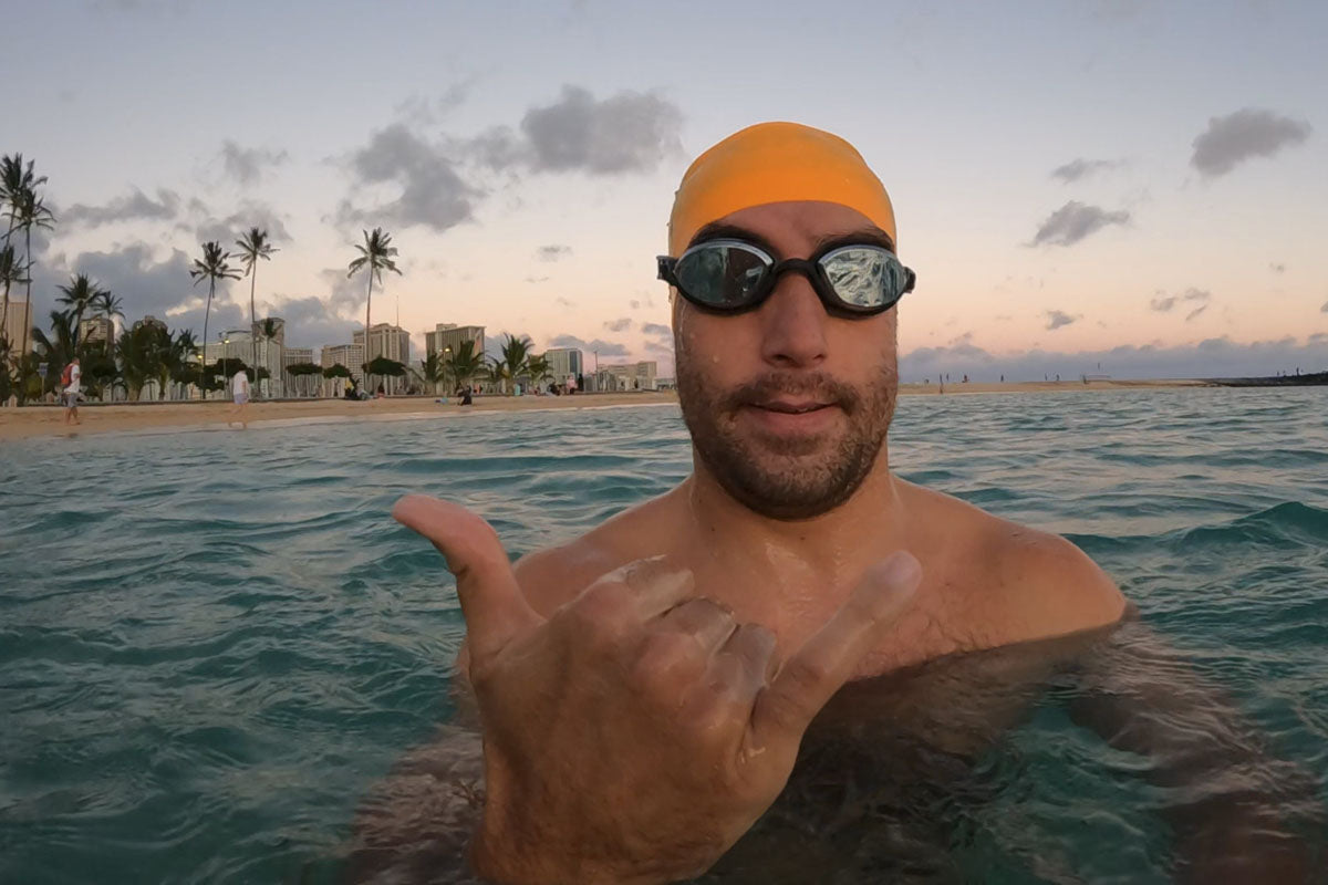 A man wearing Snake & Pig goggles and an orange swim cap makes a hang-loose sign with his hand while floating in the ocean off Waikiki Beach