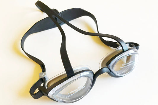 A pair of top anchor swim goggles with clear lenses and black frame and straps. A strap extends from the top of each lens over the top of the head to provide more stability.