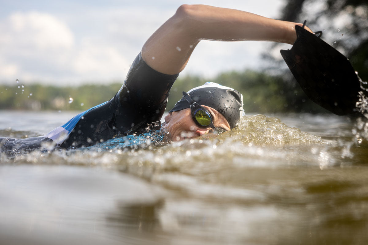 A swimrunner swims in open water with paddles. She takes a breath to the side, showing that she is wearing black tinted Junior Basilisk swim goggles.