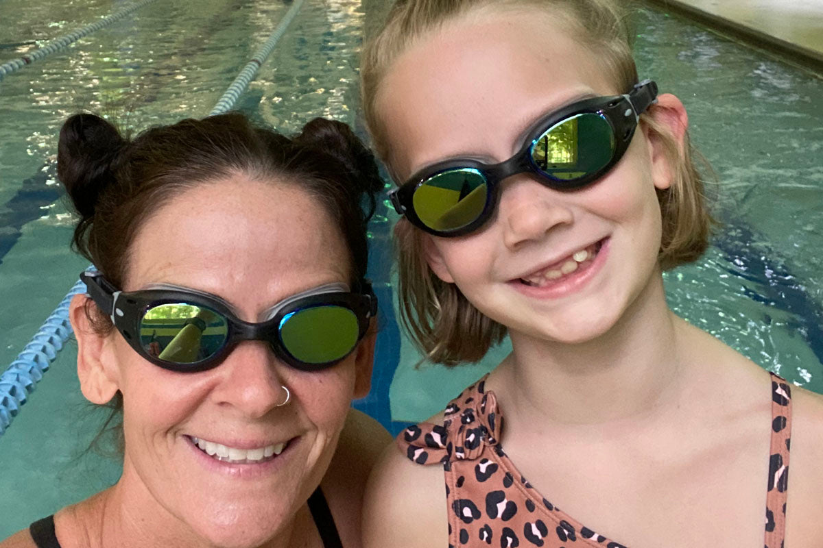 Mother and daughter swimrun enthusiasts, who both have narrow faces, wear dark tinted Junior Basilisk swim goggles at a swimming pool