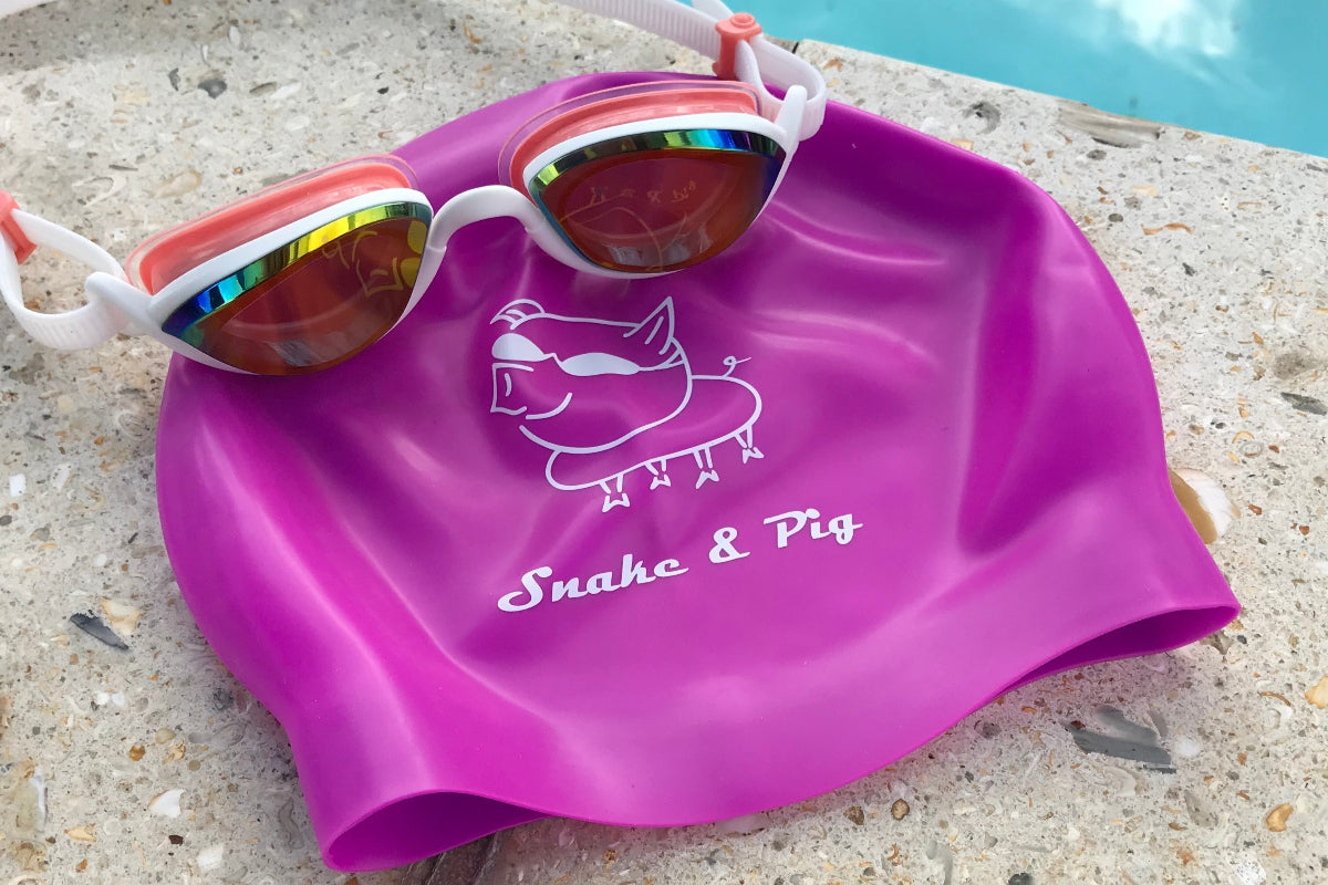 Purple silicone cap on a swimming pool deck with a pair of pink Snake & Pig goggles on top of it