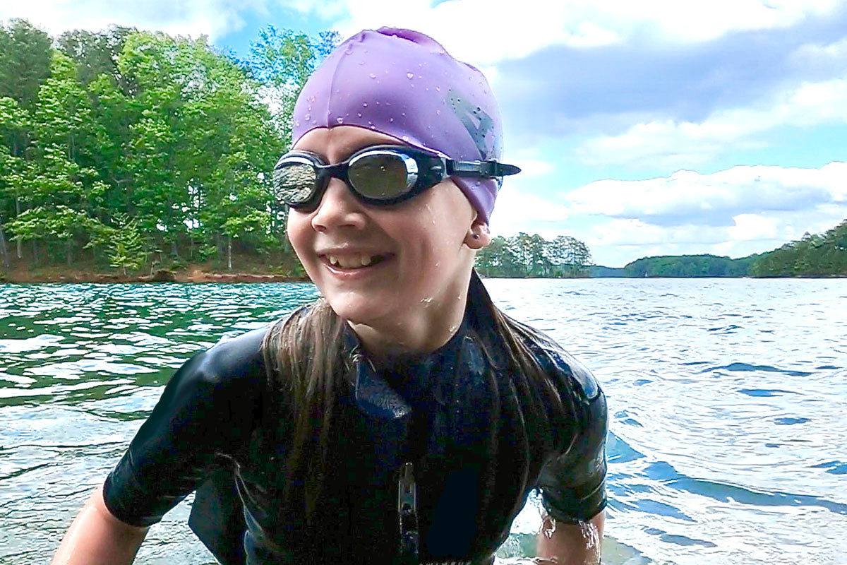 A young swimrun athlete wearing black clear junior Basilisk goggles and a purple swim cap over her long hair, prepares to exit the water. She is smiling, and her hair lays over the shoulders of her wetsuit. Green trees appear in the background.